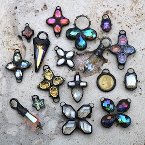 May include: A selection of iridescent crystal pendants in diverse forms, such as crosses, stars, and teardrops. The pendants are framed in dark metal and presented on a textured, light-coloured surface. Some have gold detailing.