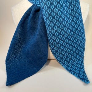 May include: A blue scarf with a diamond pattern in shades of blue. The scarf is displayed on a white mannequin. The scarf has a solid blue section and a patterned section. The scarf is made of a soft, knitted material.