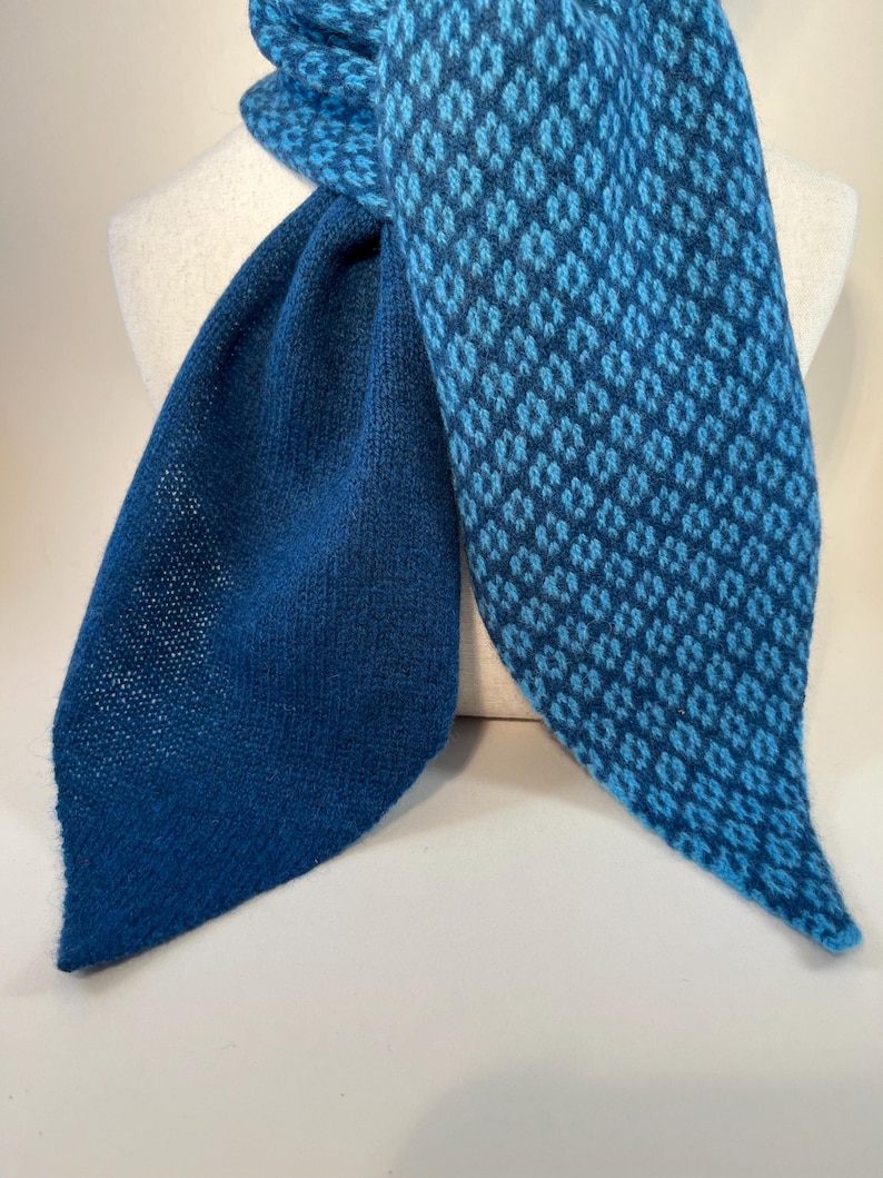 May include: A blue scarf with a diamond pattern in shades of blue. The scarf is displayed on a white mannequin. The scarf has a solid blue section and a patterned section. The scarf is made of a soft, knitted material.