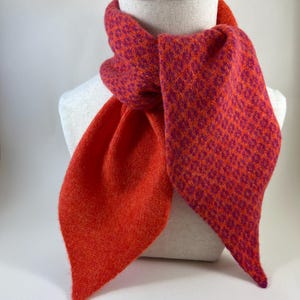 May include: A colourful scarf in orange and magenta, displayed on a mannequin. The scarf has a textured knit pattern. The ends are a solid orange, while the main body has a patterned design. The scarf is a stylish accessory.