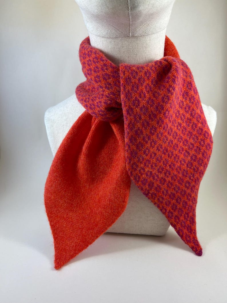May include: A colourful scarf in orange and magenta, displayed on a mannequin. The scarf has a textured knit pattern. The ends are a solid orange, while the main body has a patterned design. The scarf is a stylish accessory.