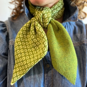 May include: A green and white patterned scarf is knotted. The scarf features a diamond pattern on one side and a solid green colour on the other. The scarf is made of a soft, knitted material.