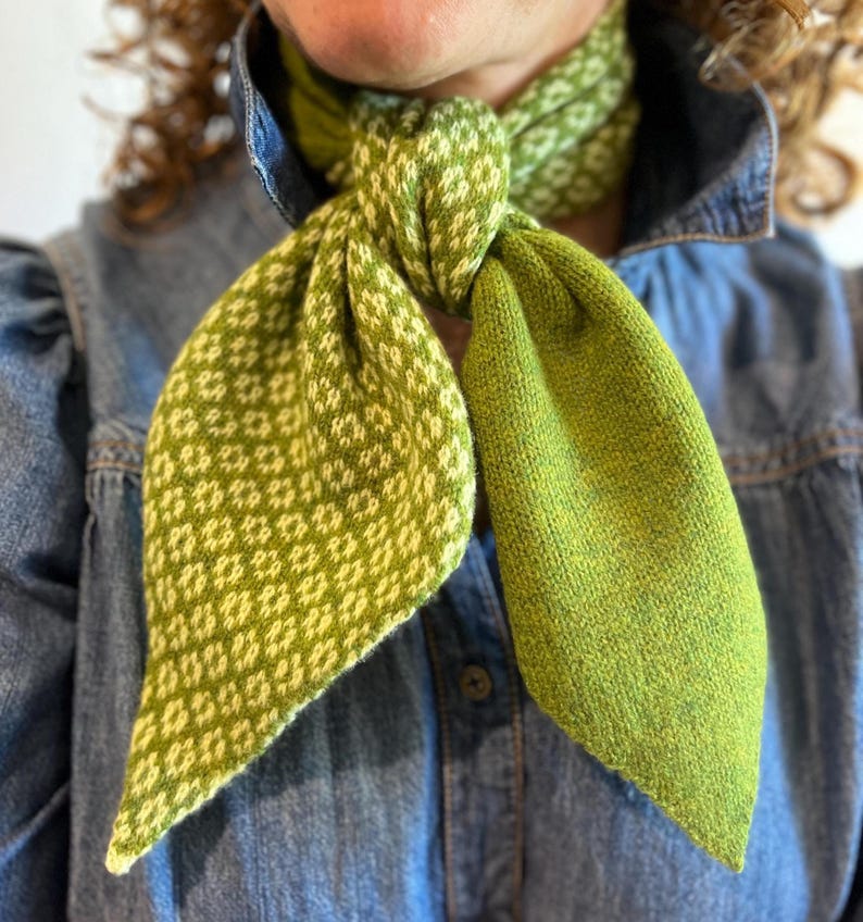 May include: A green and white patterned scarf is knotted. The scarf features a diamond pattern on one side and a solid green colour on the other. The scarf is made of a soft, knitted material.
