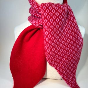 May include: A red and pink patterned scarf on a mannequin. The scarf has a solid red section and a patterned section with a repeating diamond design in pink. The scarf is made of a soft, knitted material.