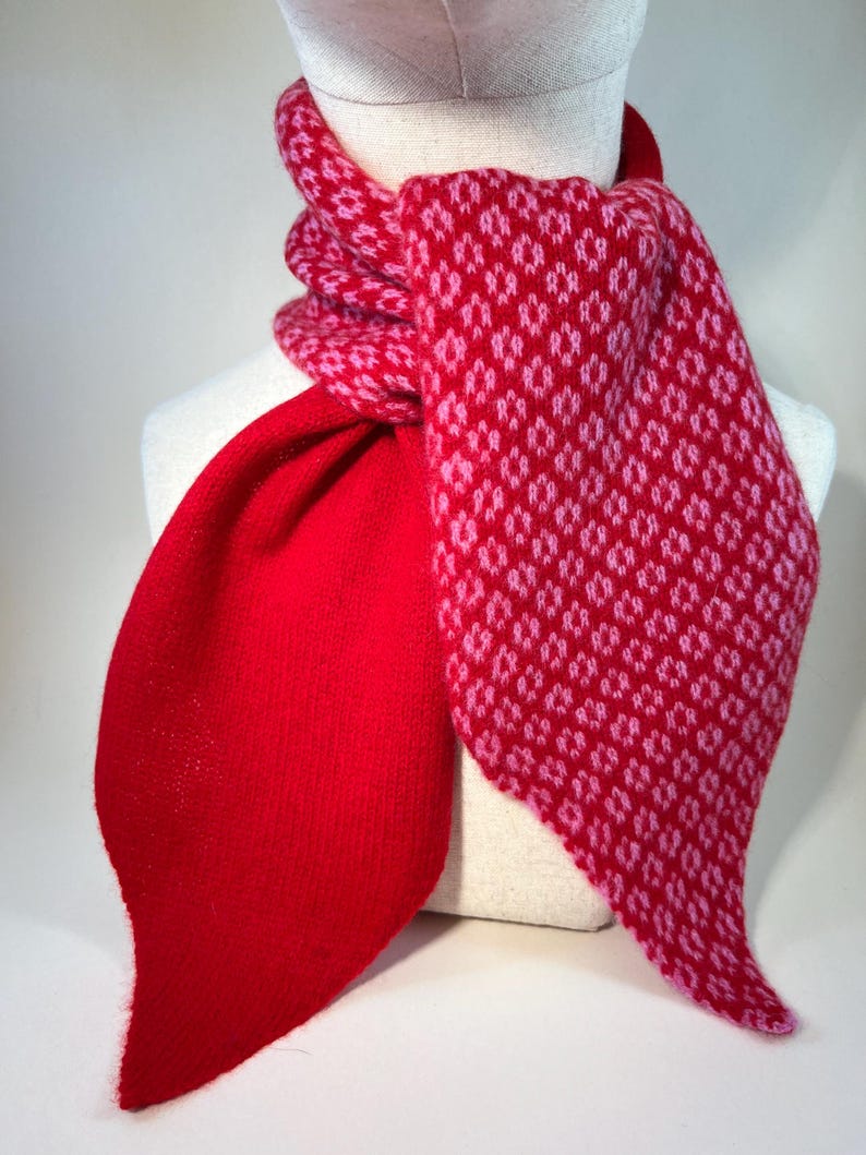 May include: A red and pink patterned scarf on a mannequin. The scarf has a solid red section and a patterned section with a repeating diamond design in pink. The scarf is made of a soft, knitted material.