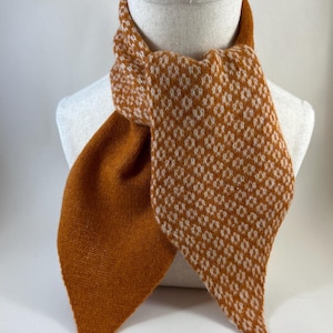 May include: A knitted scarf in shades of orange and white, displayed on a mannequin. The scarf has a diamond pattern in white on an orange base. The scarf is made of a soft, knitted material and is designed to be worn around the neck.