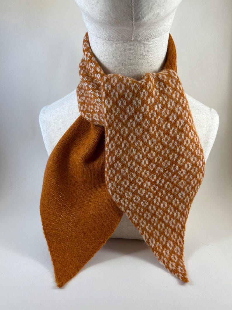 May include: A knitted scarf in shades of orange and white, displayed on a mannequin. The scarf has a diamond pattern in white on an orange base. The scarf is made of a soft, knitted material and is designed to be worn around the neck.