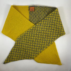 May include: Mustard yellow and grey scarf with a geometric pattern. The scarf is folded in a triangular shape, with a solid yellow section and a patterned section. The tag reads "Knits Home".
