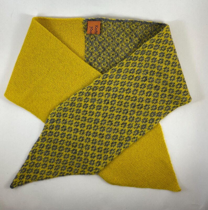 May include: Mustard yellow and grey scarf with a geometric pattern. The scarf is folded in a triangular shape, with a solid yellow section and a patterned section. The tag reads "Knits Home".