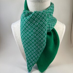 May include: A green and white patterned scarf displayed on a mannequin. The scarf has a diamond pattern on one side and a solid green colour on the other. The scarf is made of a soft, knitted material.