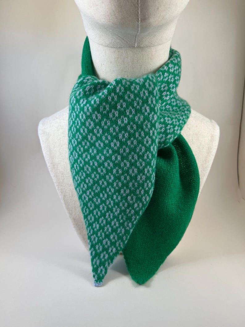 May include: A green and white patterned scarf displayed on a mannequin. The scarf has a diamond pattern on one side and a solid green colour on the other. The scarf is made of a soft, knitted material.