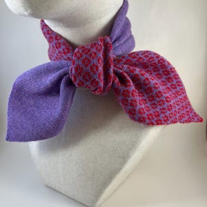 May include: A knitted scarf in shades of purple and red, displayed on a mannequin head. The scarf is tied in a bow, with a solid purple section at the neck and a patterned red and purple section forming the bow.