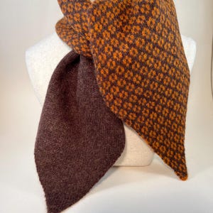 May include: A brown and orange scarf with a geometric pattern. The scarf is draped over a mannequin. The scarf has a solid brown section and a patterned section with orange diamonds on a brown background. The scarf is made of a soft, textured material.