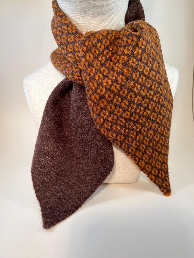 May include: A brown and orange scarf with a geometric pattern. The scarf is draped over a mannequin. The scarf has a solid brown section and a patterned section with orange diamonds on a brown background. The scarf is made of a soft, textured material.