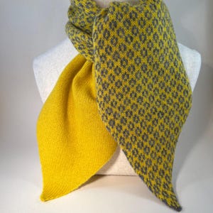May include: A yellow and grey knitted scarf displayed on a mannequin. The scarf has a geometric pattern in grey on one side and a solid yellow colour on the other. The scarf is made of soft material.