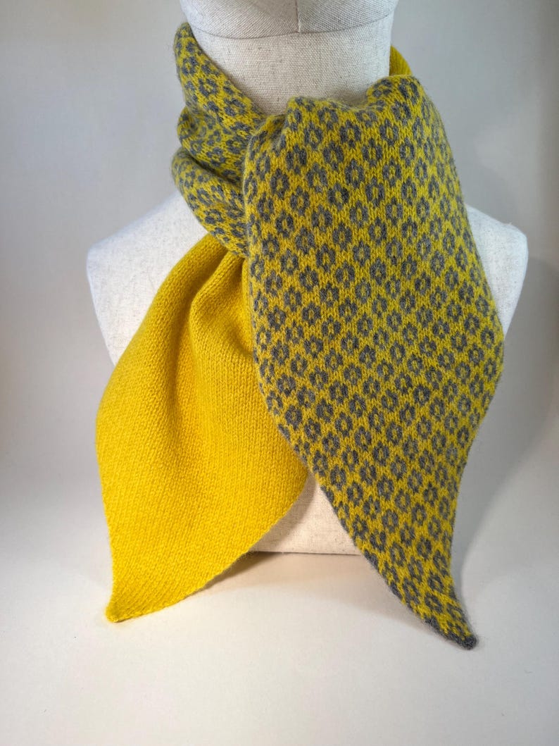 May include: A yellow and grey knitted scarf displayed on a mannequin. The scarf has a geometric pattern in grey on one side and a solid yellow colour on the other. The scarf is made of soft material.