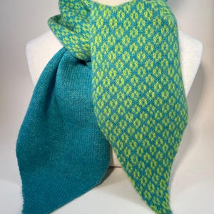 May include: A teal and lime green knitted scarf. The scarf has a diamond pattern in lime green on a teal background. The scarf is draped over a mannequin, highlighting its design and texture. The scarf's ends are pointed.