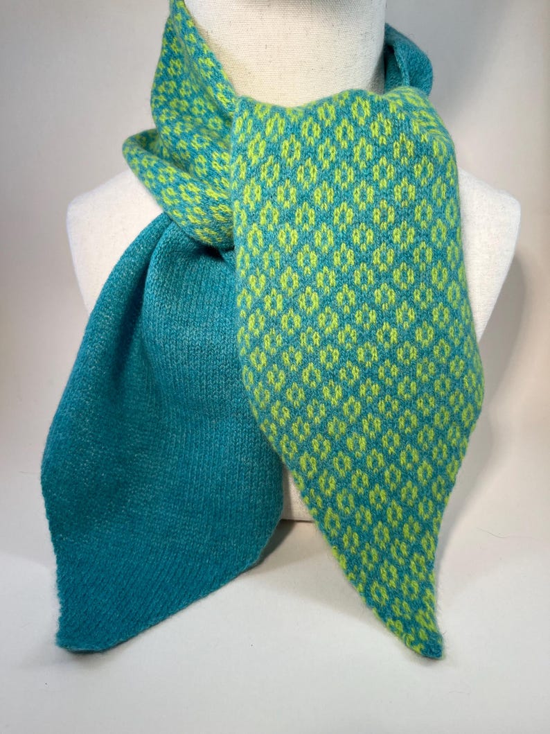 May include: A teal and lime green knitted scarf. The scarf has a diamond pattern in lime green on a teal background. The scarf is draped over a mannequin, highlighting its design and texture. The scarf's ends are pointed.
