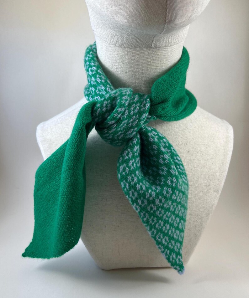 May include: A green and white patterned scarf is displayed on a mannequin. The scarf is knotted at the front, with the ends falling down. The pattern features small white designs on a green backdrop. The scarf is made of a soft, textured material, suitable for wear.