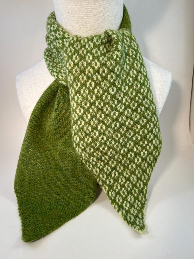 May include: A green and white patterned scarf displayed on a mannequin. The scarf has a diamond pattern in white against a green background. The scarf is made of a soft, knitted material and is designed to be worn around the neck.