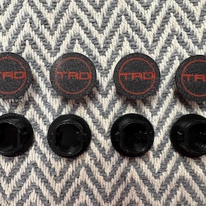 May include: A set of four circular black valve stem caps featuring the red TRD logo enclosed in a red circle on the top surface. The underside of each cap is black with a hollow cylindrical shape and internal threading for secure attachment.