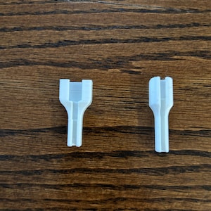 May include: Two white, 3D-printed plastic connectors. One connector has a U-shaped top with a rectangular opening, and the other has a rounded top. Both connectors have a cylindrical base and are set against a wood grain background.