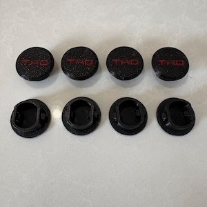 以下が含まれることがあります： 赤色の「TRD」の文字が入った黒い円形のオブジェクトが8つ。4つは文字が書かれた平らな上面を持ち、4つは底面に凹んだデザインがあります。オブジェクトは2列4つに配置されています。