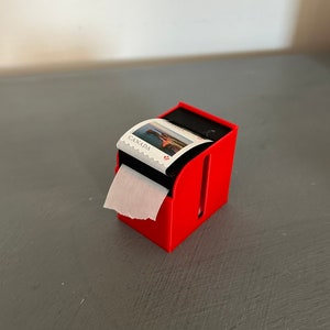 May include: A red plastic dispenser with a roll of Canadian stamps. The stamps feature a picture of a lighthouse and the word "CANADA".