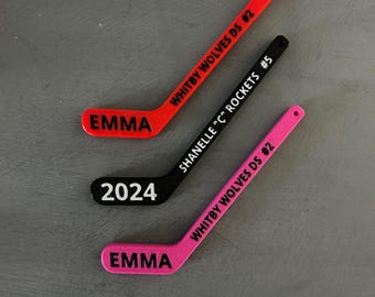 May include: Three miniature hockey sticks in red, black, and pink. Each stick has text: "EMMA", "2024", "Shanelle 'C' Rockets #5", and "Whitby Wolves DS #2". The sticks are likely personalized sports-themed items.