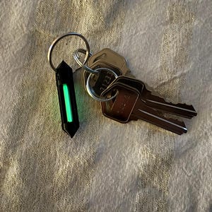 Puede incluir: Un llavero negro de forma geométrica con una barra central verde brillante, sujeto a un llavero plateado. Varias llaves de metal también están presentes, apoyadas sobre una superficie texturizada.