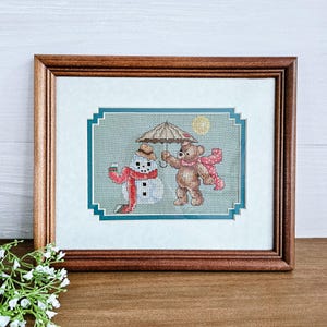 May include: A framed cross-stitch embroidery featuring a brown bear holding an umbrella over a snowman. The bear is wearing a red scarf and the snowman is wearing a red scarf and hat. The embroidery is set against a green background with a blue border.