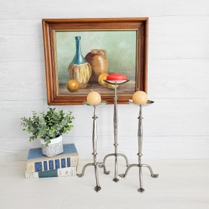 May include: Three silver candle holders with a tripod base and a round top. Each holder has a white candle on top. The candle holders are in front of a framed painting of a still life with a bottle, a jug, and fruit.