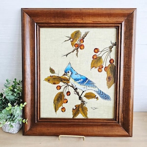 May include: Framed artwork featuring a blue jay perched on a branch with berries and autumn leaves. The bird is painted in shades of blue and white, set against a neutral background. The wooden frame adds a rustic touch.