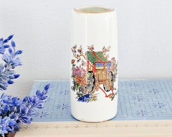 May include: White ceramic vase with a floral design featuring a Japanese-style cart with a red rope. The cart is pulled by a horse and has a green and blue roof. The vase has a gold rim.