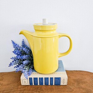 May include: A yellow ceramic coffee pot with a white lid and a handle. The pot is sitting on a stack of books with blue and white covers.