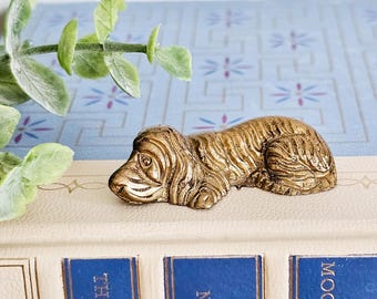 Op de afbeelding: Een gouden metalen beeldje van een liggende hond. Het beeldje is gedetailleerd met textuur in de vacht en een realistische uitdrukking.