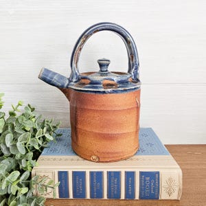 May include: A handmade ceramic teapot with a brown body and a blue handle, spout, and rim. The teapot is sitting on top of a stack of books. The books have blue covers and gold lettering. The teapot is approximately 6 inches tall.