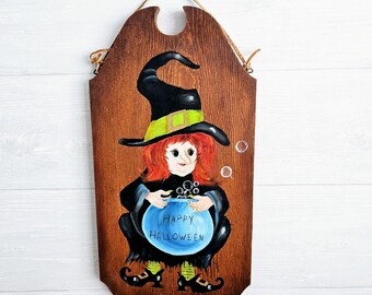 Op de afbeelding: Een houten bord met een geschilderd beeld van een heks die een blauwe ketel vasthoudt waarop "Happy Halloween" staat. De heks heeft rood haar, een zwarte hoed met een groene band en draagt een zwarte jurk. Het bord heeft een bruine houtnerf en hangt aan een touw.