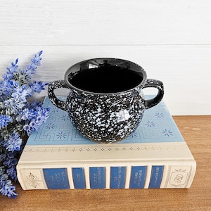 May include: A black and white speckled ceramic bowl with two handles. The bowl is sitting on a light blue book with a floral design. The bowl has a glossy finish and is filled with a dark liquid.