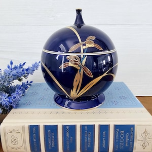 May include: A round blue and gold ceramic jar with a lid. The jar is decorated with a floral design of gold and brown flowers. The jar is sitting on a stack of books.