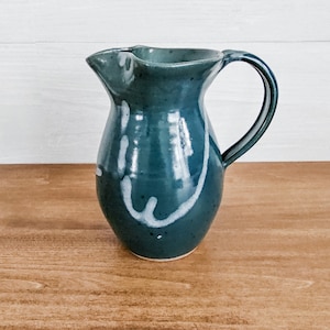 May include: A teal blue ceramic pitcher with white glaze accents. The pitcher has a rounded body and a handle.