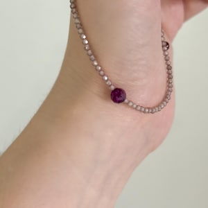 May include: A delicate bracelet featuring small, faceted beads in a pale, neutral color. A larger, round, deep red bead is centered on the bracelet. The bracelet is worn on a wrist.