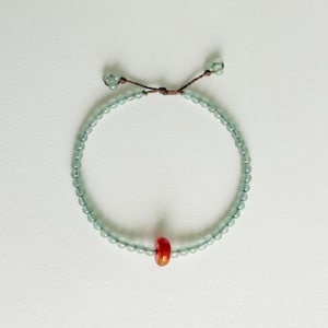 Op de afbeelding: Een delicate armband met kleine, lichtblauwe kralen. Een enkele, helderrode kraal bevindt zich in het midden. De armband is vastgemaakt met een bruin koord, waardoor hij verstelbaar is. Sieraden.