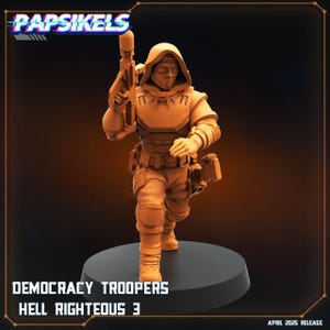 May include: A tan-colored figurine of a futuristic soldier, holding a rifle and wearing a hooded uniform with tactical gear. The text "Papsikels" is at the top, and "DEMOCRACY TROOPERS HELL RIGHTEOUS 3" is at the bottom. "APRIL 2025 RELEASE" is also visible.
