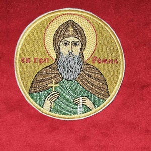 Op de afbeelding: Cirkelvormige geborduurde patch met een religieuze figuur met een halo, die een kruis en een rozenkrans vasthoudt. De figuur heeft een lange baard en draagt een bruine kap en een groen gewaad. De patch heeft gouden en rode accenten en tekst in een vreemde taal.