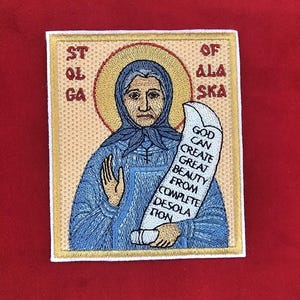 Op de afbeelding: Geborduurde patch met een religieus icoon van St. Olga van Alaska. De afbeelding toont een vrouw in blauwe gewaden met een halo, die een rol vasthoudt met de tekst "GOD CAN CREATE GREAT BEAUTY FROM COMPLETE DESOLATION". De patch heeft een gouden rand.