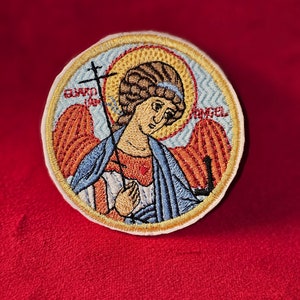 Op de afbeelding: Geborduurde patch met een kleurrijke afbeelding van een engel met vleugels, die een kruis vasthoudt. De engel draagt een blauwe robe en heeft een halo. De patch heeft een witte rand en de woorden "BESCHERMENDE ENGEL" zijn rond de rand geborduurd.