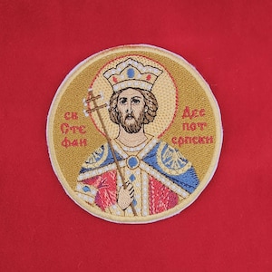 May include: Embroidered patch featuring a religious icon with a halo, crown, and staff. The patch has gold, red, blue, and white details. The text in the image is in a foreign language. The patch is circular and set against a red background.