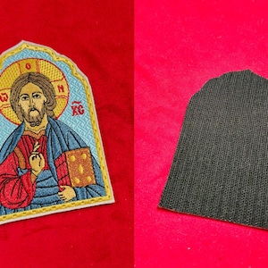 Op de afbeelding: Geborduurde strijkpatch met een religieus icoon van Jezus Christus. De patch heeft een blauwe achtergrond en gouden rand en bevindt zich op een rood oppervlak.