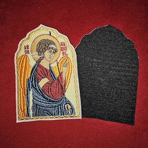 May include: Embroidered patch featuring a colorful depiction of an angel with wings, holding a staff. The patch is surrounded by a gold frame and has the text "ARCHANGEL" in the top corners. The patch is attached to a black fabric backing.
